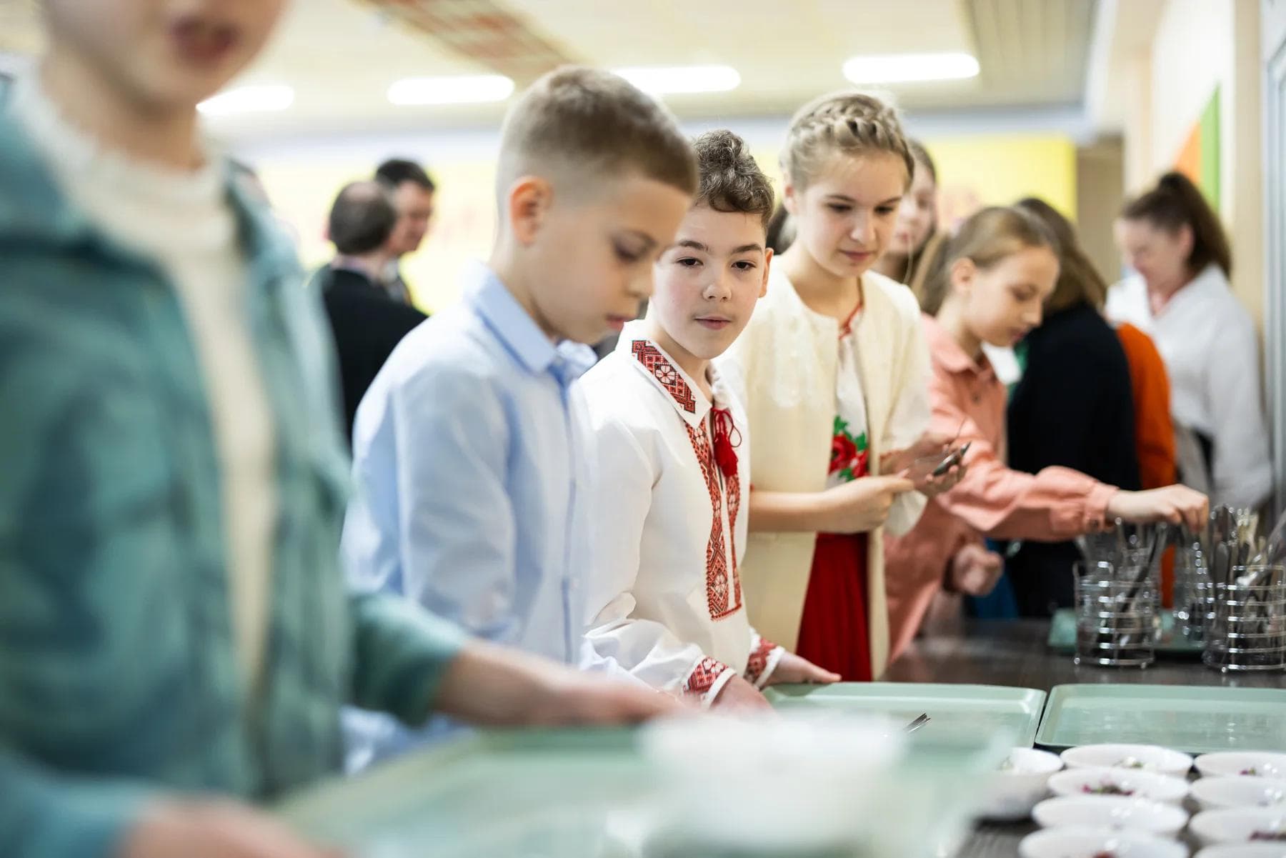 Діти стоять у черзі в шкільній їдальні з тацями та приборами в руках, готуючись отримати їжу. Деякі з них одягнені в традиційний вишитий одяг.